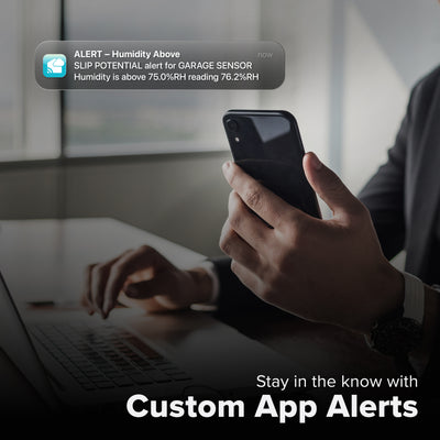 La Crosse Technology Alert and Monitoring system showing a male sitting at a desk on a laptop computer looking at his smartphone receiving an alert that the humidity level is high in the garage outside of the parameters set for the temperature and humidity sensor with text that says stay in the know with Custom Alerts