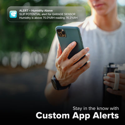 La Crosse Technology Alert and Monitoring system showing a female with a travel mug looking at her smartphone receiving an alert that the humidity level is high in the garage outside of the parameters set for the temperature and humidity sensor with text that says stay in the know with Custom Alerts