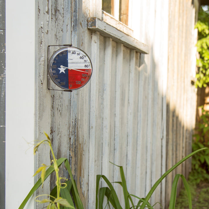 Lifestyle image of the 104-105TX 5-inch Dial Thermometer with Bracket that is positioned on the left side mounted to a whitewashed, weathered board fence. The dial features black numbers on a rustic Americana flag in red, white, and blue with a white star, over rustic weathered boards, surrounded by a black frame with an adjustable bracket for mounting. Featuring a silver pointer that provides the temperature reading in Fahrenheit from -60-120 degrees.