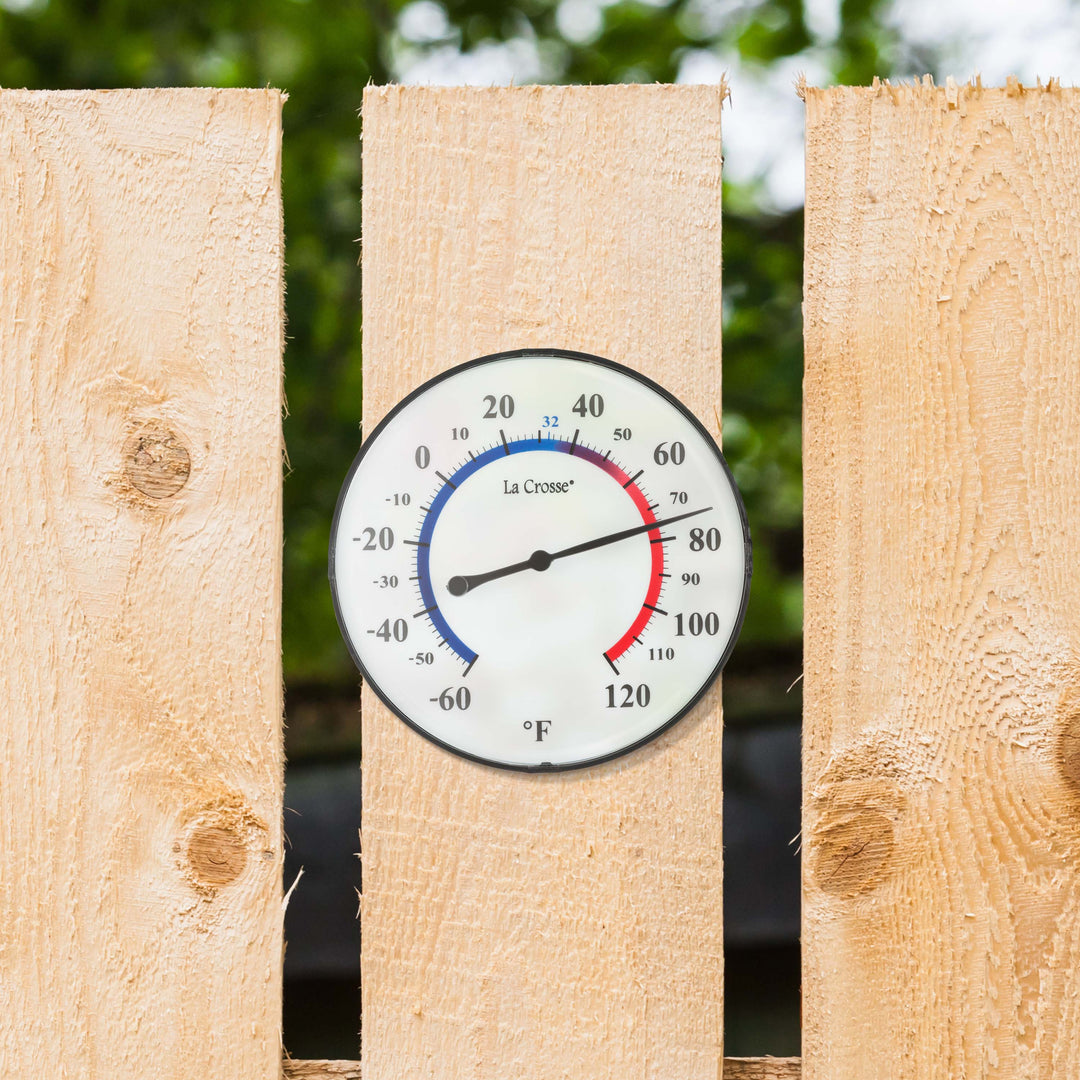 Lifestyle image of the  104-105 5-inch Dial Thermometer with Bracket for multiple mounting options. Hang using the bracket in four different positions, or as a kickstand for table placement. The dial reads in degrees Fahrenheit from -60 to 120, with a color scale from blue (cold) to red (warm). The thermometer is mounted on a light wood fence with hints of green trees visible through the slats.
