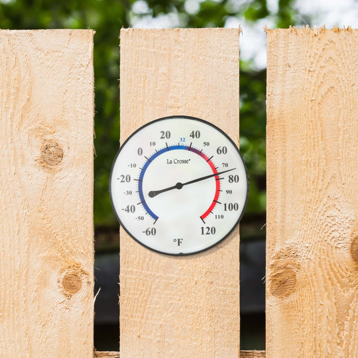 Lifestyle image of the  104-105 5-inch Dial Thermometer with Bracket for multiple mounting options. Hang using the bracket in four different positions, or as a kickstand for table placement. The dial reads in degrees Fahrenheit from -60 to 120, with a color scale from blue (cold) to red (warm). The thermometer is mounted on a light wood fence with hints of green trees visible through the slats.