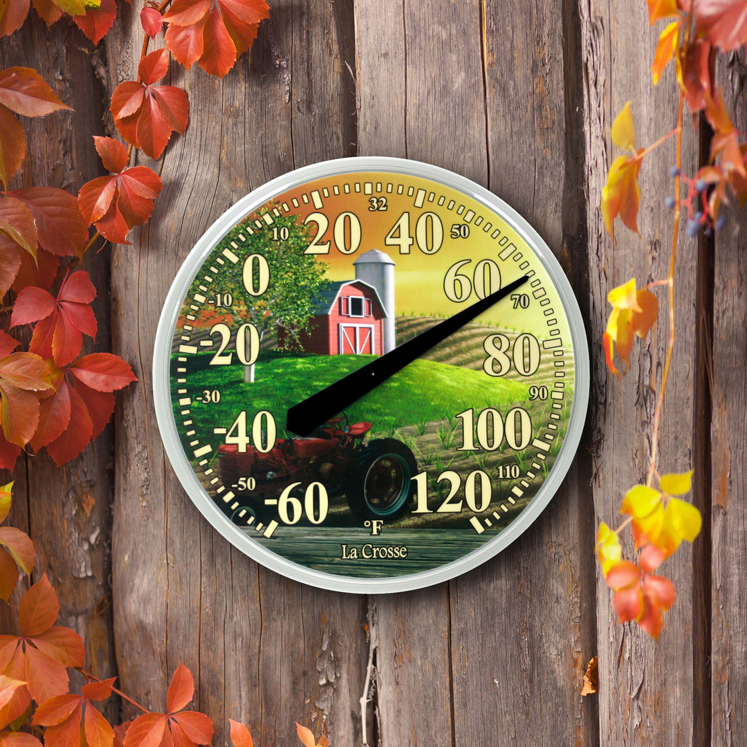Lifestyle image of the 104-114-FARM 13.5-inch Dial Thermometer - Barn, with key hider, temperature scale in Fahrenheit -60 to 120 degrees with a red-tipped pointer in a white slimline case hanging on a reclaimed barnwood fence with fall foliage