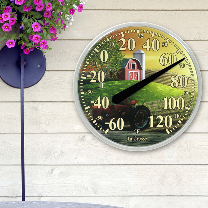Lifestyle image of the 104-114-FARM 13.5-inch Dial Thermometer - Barn, with key hider, temperature scale in Fahrenheit -60 to 120 degrees with a red-tipped pointer in a white slimline case hanging on a white wood siding house next to pink flowers