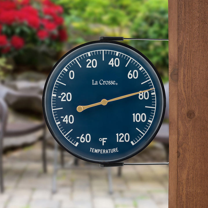 Lifestyle image of the 104-1512E 5-inch Dial Thermometer with Bracket features a navy blue dial with white temperature readings in a Fahrenheit scale from -60 to 120 degrees, with an antique gold pointer shown mounted on a post in a backyard paver patio with trees and a red flowering bush in the background