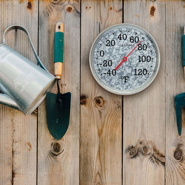 Lifestyle image of the 104-2822 8-inch Galvanized Dial Thermometer, galvanized metal dial with black number markings and a red pointer with a temperature scale from -40 to 120 degrees Fahrenheit hanging on a wood fence with garden tools