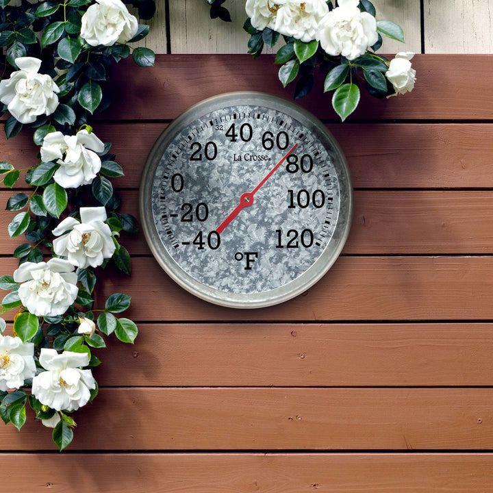 104-2822 8-inch Galvanized Dial Thermometer, galvanized metal dial with black number markings and a red pointer with a temperature scale from -40 to 120 degrees Fahrenheit, hanging on a brown painted fence with a white flower vine