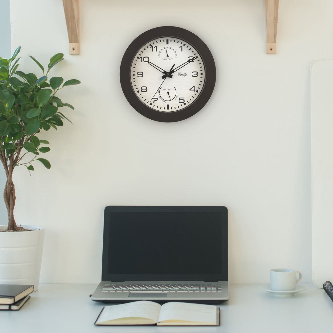 29005 10-inch Waylon indoor outdoor wall clock with temperature and humidity, linen design dial with dark brown frame features stylized cut out hands: hour and minute, and a second hand hanging above a desk with a laptop computer, book, plant and coffee cup