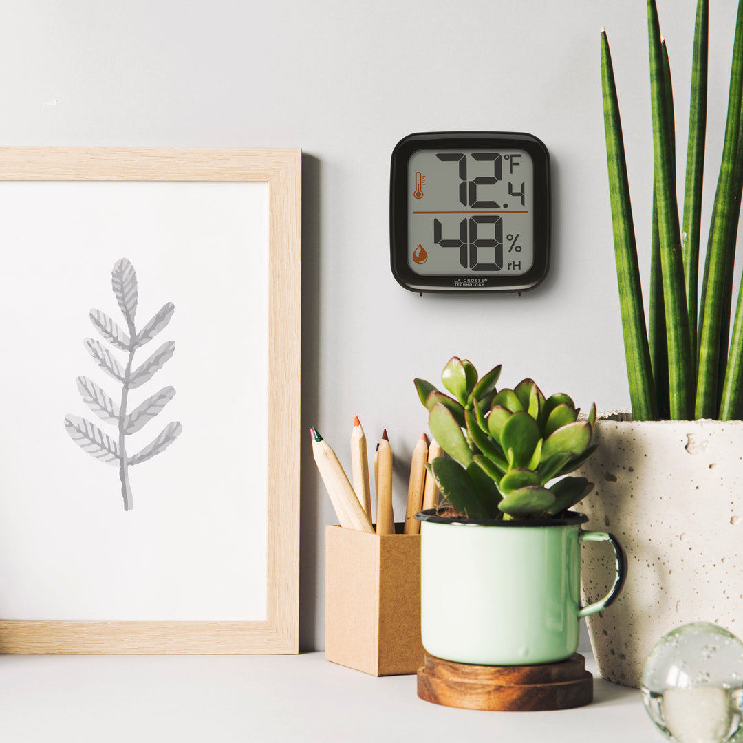 Lifestyle image of the 302-1407B Thermometer with Temperature and Humidity - Black hanging on a light gray wall above a desk. The desk has plants, a pencil cup an organic piece of artwork. The thermometer features a black square case and a digital screen with large temperature and humidity readings. Next to the digits are corresponding temperature and humidity icons in orange.