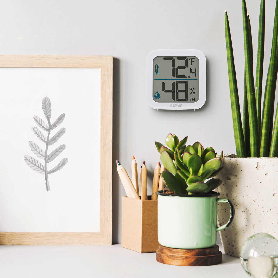 Lifestyle image of the 302-1407W Thermometer with Temperature and Humidity - White hanging on a light gray wall above a desk. The desk has plants, a pencil cup an organic piece of artwork. The thermometer features a white square case and a digital screen with large temperature and humidity readings. There are temperature and humidity icons in blue next to their corresponding readings.