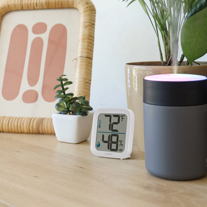 Lifestyle image of the 302-1407W Thermometer with Temperature and Humidity - White. The thermometer is shown sitting on a light-colored wood shelf or table with plants, a picture frame, and a light-up humidifier. The thermometer features a white square case and a digital screen with large temperature and humidity readings. There are temperature and humidity icons in blue next to their corresponding readings.