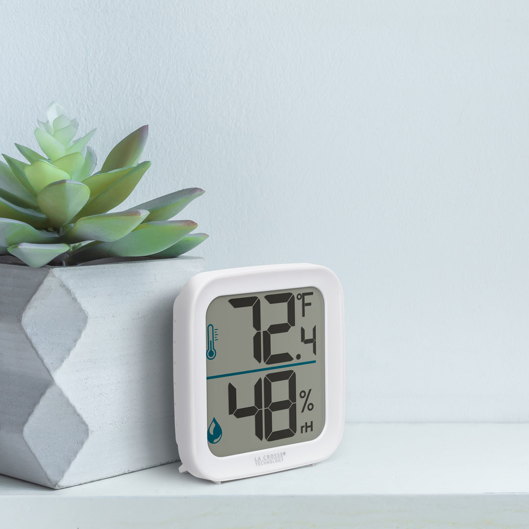 Lifestyle image of the 302-1407W Thermometer with Temperature and Humidity - White sitting on a white shelf next to a whitewashed wood vase with succulents. The thermometer features a white square case and a digital screen with large temperature and humidity readings. Next to the digits are corresponding temperature and humidity icons in blue.