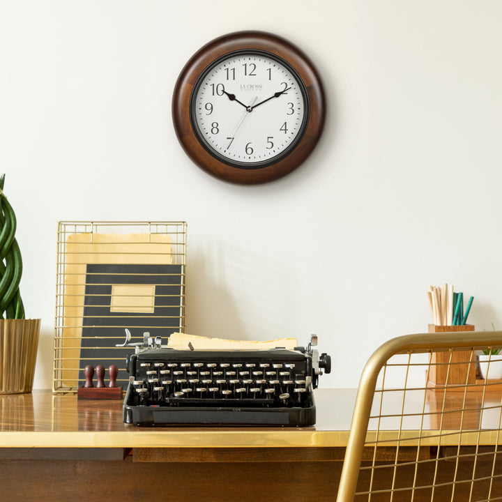 Lifestyle image of a desk with a vintage typewriter, gold mesh chair, and desk accessories with the 404-2625 Linwood wall clock hanging on a cream colored wall above the desk, the clock features a dark stain wood frame surrounding a white dial with simple black numerals and time markings, black metal hour, minute, and second hands.