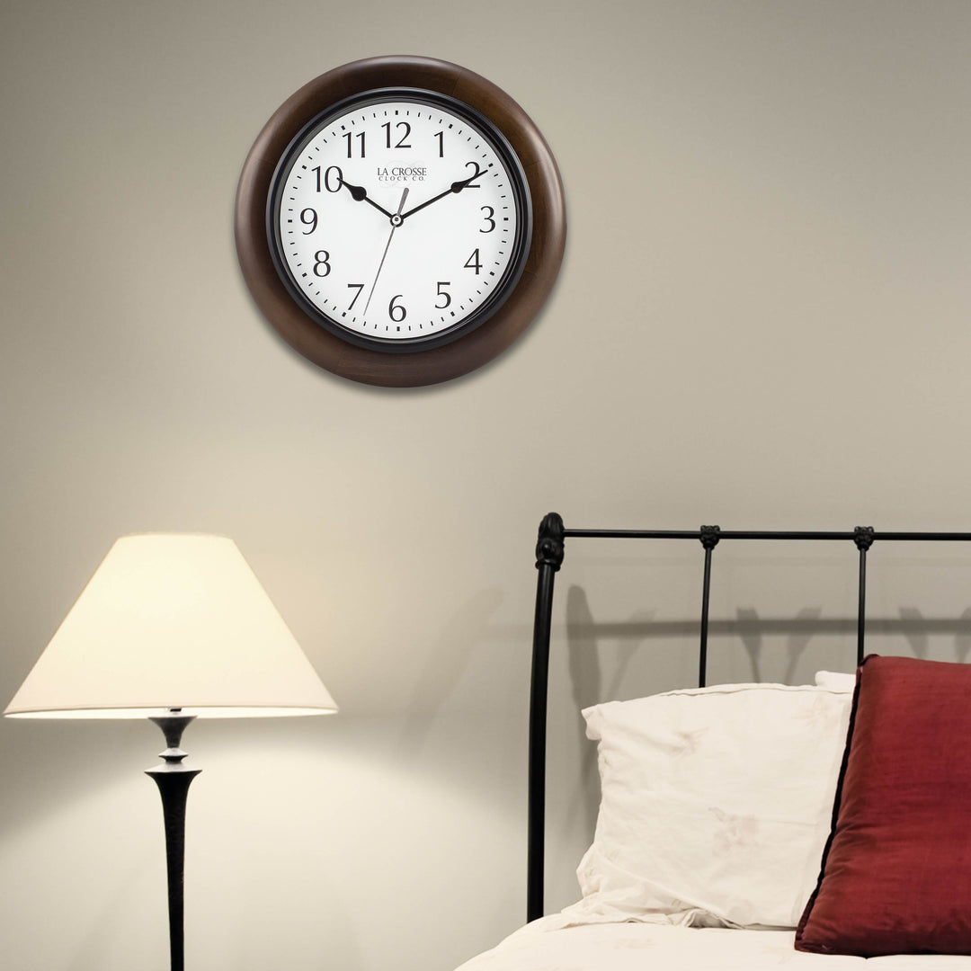 404-2625 Linwood wall clock features a dark stained wood frame surrounding a white dial with simple black numerals and time markings, black metal hour and minute hands, and a silver second hand hanging on a tan wall above a black metal framed bed with cream bedding and a red accent pillow