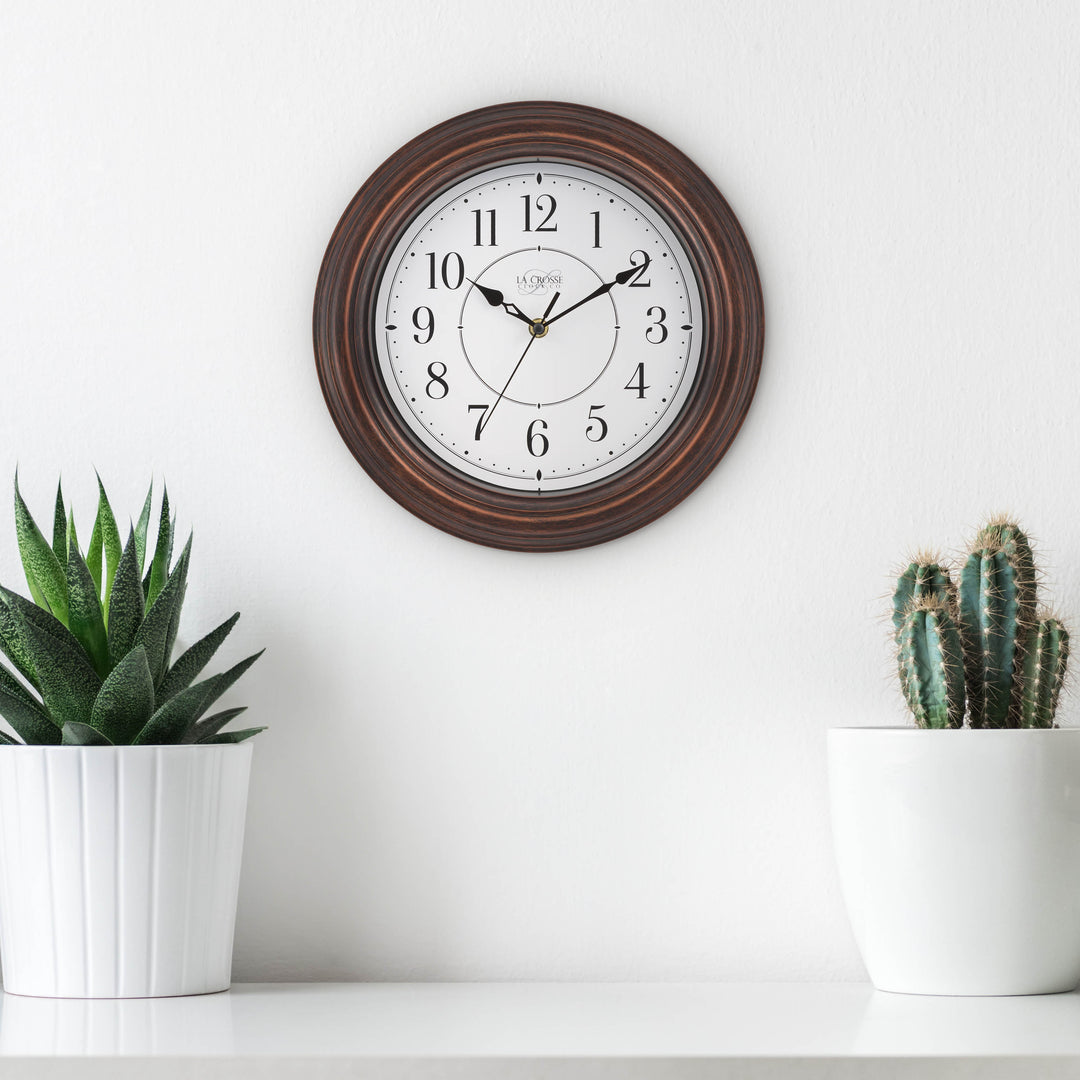 Lifestyle image of the 404-2630W Evelyn wall clock in bronze with a white dial and black stylized Arabic numbers hanging white wall above a white shelf with two white potted plants, cactus an succelent