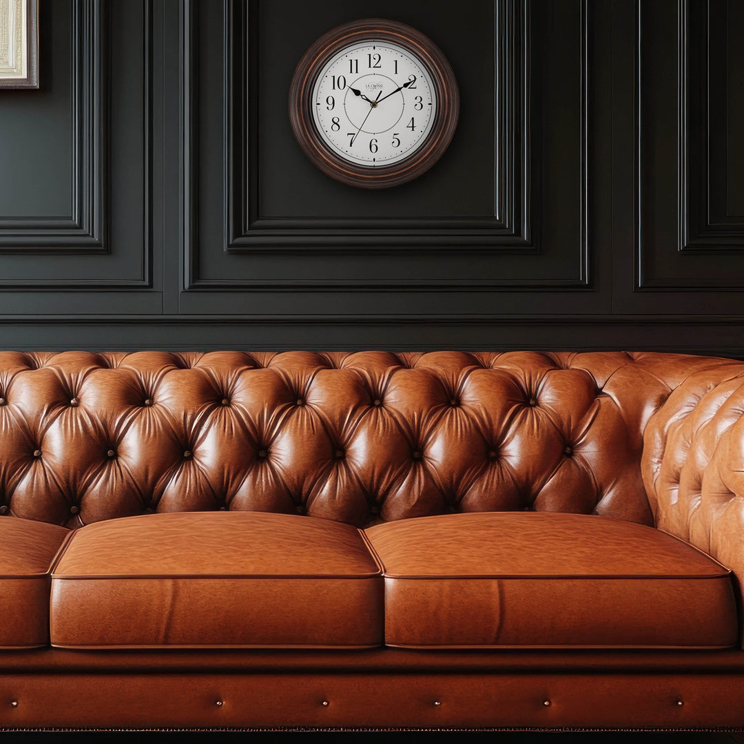 Lifestyle image with a camel leather tufted couch inf front of a dark gray wall with molding with the 404-2630W Evelyn wall clock in bronze with a white dial and black stylized Arabic numbers hanging above the couch