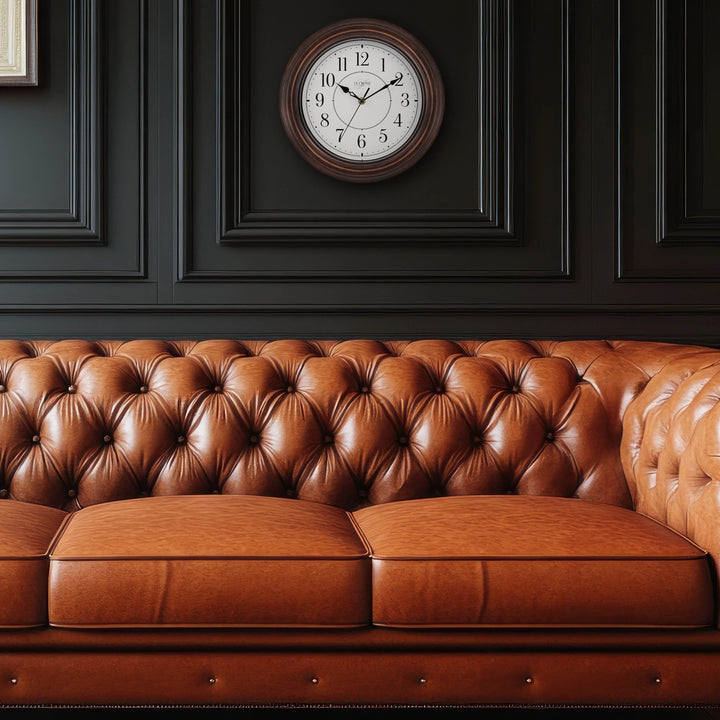 Lifestyle image with a camel leather tufted couch inf front of a dark gray wall with molding with the 404-2630W Evelyn wall clock in bronze with a white dial and black stylized Arabic numbers hanging above the couch
