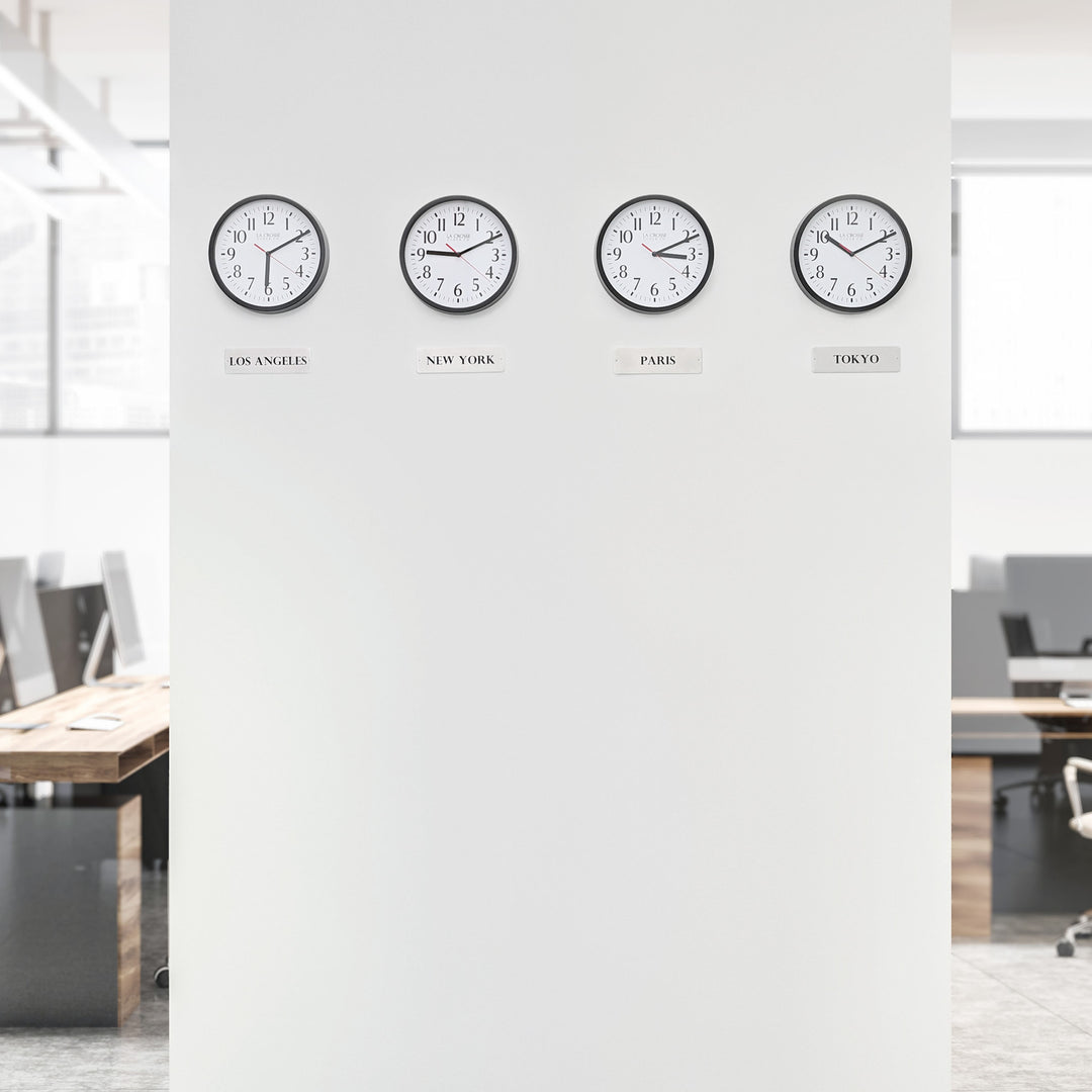 Lifestyle image of the 404-3020B-4TZ Four Pack 8-inch Basic Wall Clock with City Plaques shown hanging on a white wall with office cubicles and desks in the background. The clocks feature black frames with white clock faces, black numbers, black hour and minute hands, and red second hands.