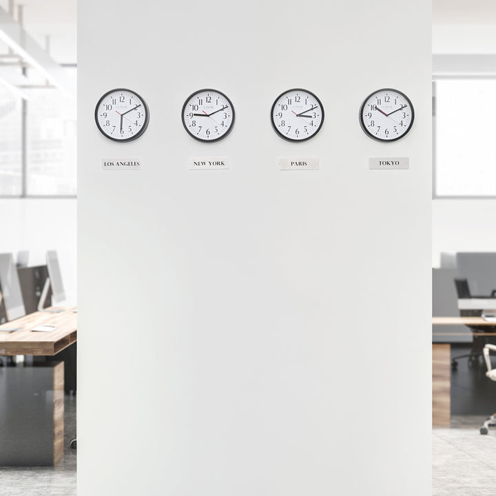 Lifestyle image of the 404-3020B-4TZ Four Pack 8-inch Basic Wall Clock with City Plaques shown hanging on a white wall with office cubicles and desks in the background. The clocks feature black frames with white clock faces, black numbers, black hour and minute hands, and red second hands.