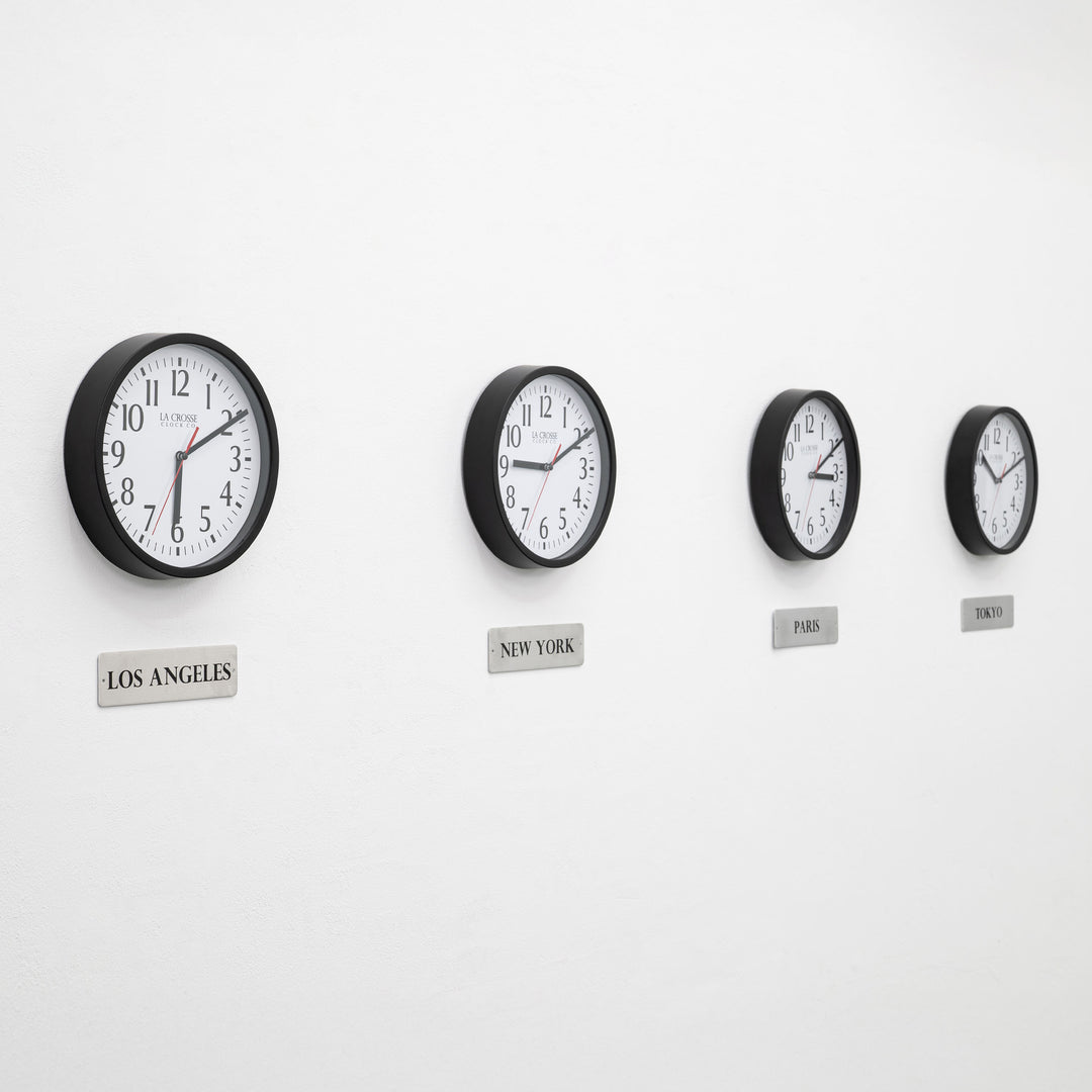 Lifestyle image of the 404-3020B-4TZ Four Pack 8-inch Basic Wall Clock with City Plaques. Four black 8-inch round wall clocks with white faces and black numerals are mounted in a horizontal row on a light gray wall. Each clock has a red second hand and displays a different time. Beneath each clock is a small rectangular plaque labeling a city: “Los Angeles,” “New York,” “Paris,” and “Tokyo,” indicating different time zones.