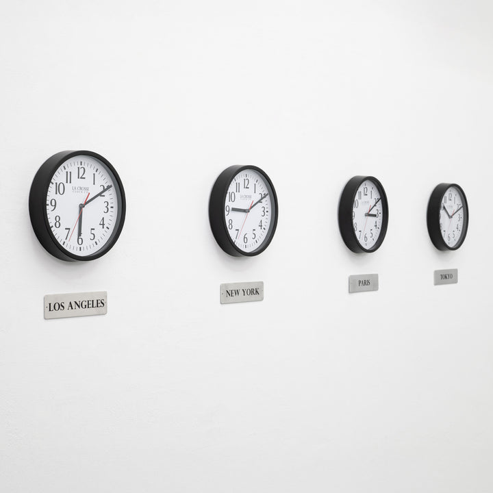 Lifestyle image of the 404-3020B-4TZ Four Pack 8-inch Basic Wall Clock with City Plaques. Four black 8-inch round wall clocks with white faces and black numerals are mounted in a horizontal row on a light gray wall. Each clock has a red second hand and displays a different time. Beneath each clock is a small rectangular plaque labeling a city: “Los Angeles,” “New York,” “Paris,” and “Tokyo,” indicating different time zones.