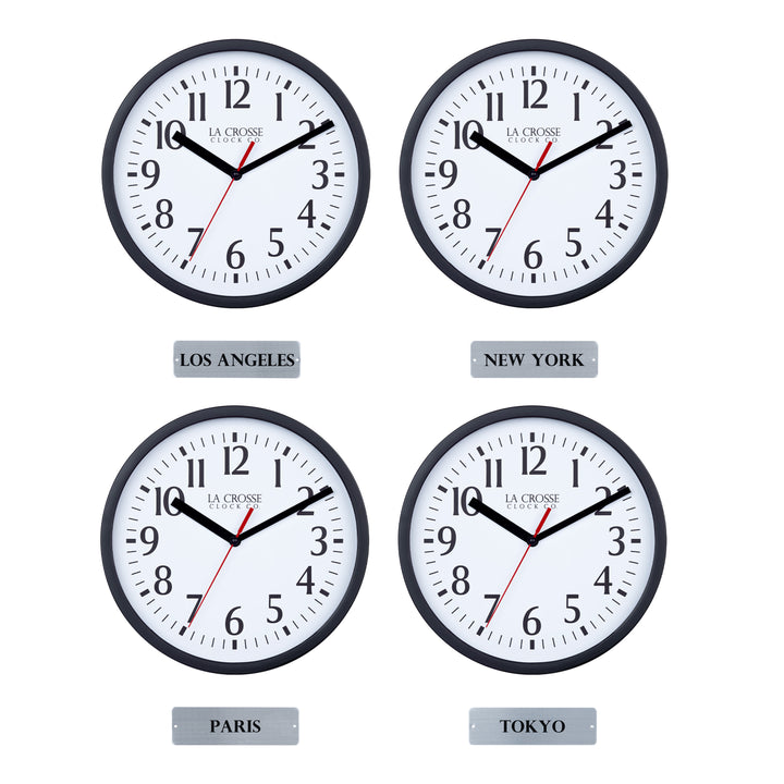 Head-on image of the 404-3020B-4TZ Four Pack 8-inch Basic Wall Clock with City Plaques showing all four clocks. The clocks feature black frames with white dials, black numbers, black hour and minute hands, and a red second hand. Beneath each clock is a small rectangular plaque labeling a city: “Los Angeles,” “New York,” “Paris,” and “Tokyo,” indicating different time zones.