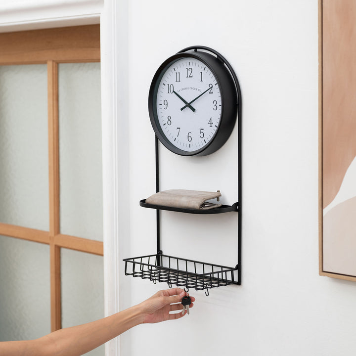 Lifestyle image of the 404-3022 Depot Wall Clock with Basket and Shelf hanging in an entryway showing a wallet on the shelf, and someone hanging keys on the "S" hook. The clock features a Black Metal Frame with a shelf and a basket, an archival paper-inspired dial, black numbers, and black metal hour and minute hands.