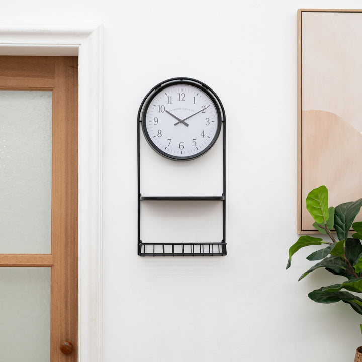 Lifestyle image of the 404-3022 Depot Wall Clock with Basket and Shelf hanging in an entryway with a plant and picture. The clock features a Black Metal Frame with a shelf and a basket, an archival paper-inspired dial, black numbers, and black metal hour and minute hands.