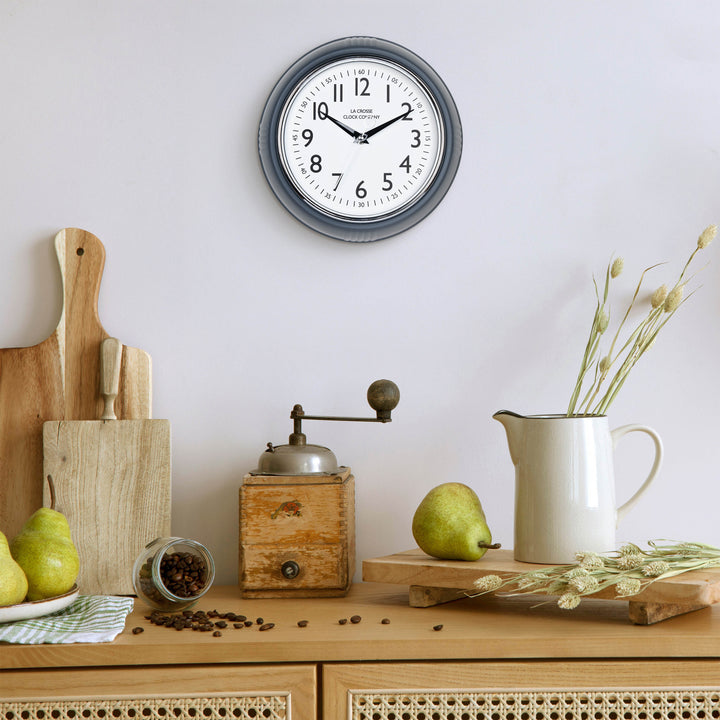 Lifestyle image of the 404-3024DS 9.5-inch Diner Clock – Dark Slate shown hanging on a light grey wall above a wood side table with an antique coffee grinder, a white ceramic pitcher, pears, coffee, and other kitchen items. The clock features a retro-style dark slate frame with a white dial, chrome bezel, black numbers and minute markings, black metal hands, and a silver second hand.