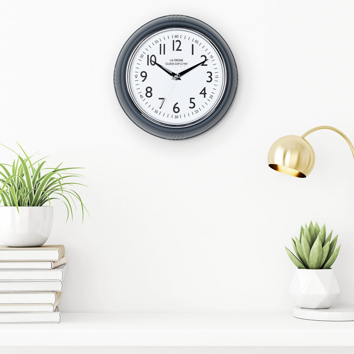 Lifestyle image of the 404-3024DS 9.5-inch Diner Clock – Dark Slate shown hanging on a white wall above a white shelf with plants, books, and a retro lamp. The clock features a retro-style dark slate frame with a white dial, chrome bezel, black numbers and minute markings, black metal hands, and a silver second hand.