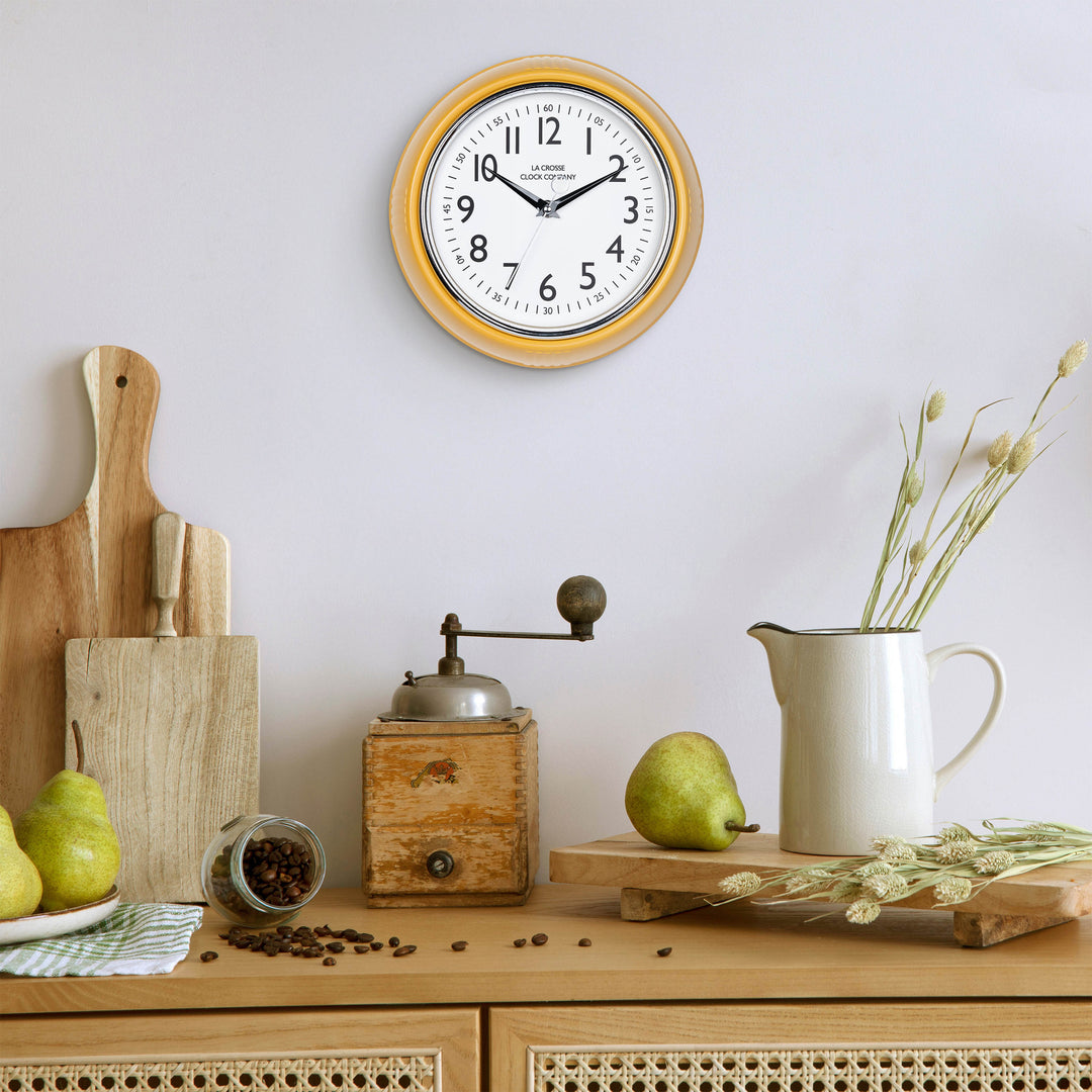 Lifestyle image of the 404-3024MY 9.5-inch Diner Clock – Mustard Yellow shown hanging on a light grey wall above a wood side table with an antique coffee grinder, a white ceramic pitcher, pears, coffee, and other kitchen items. The clock features a retro-style mustard yellow frame with a white dial, chrome bezel, black numbers and minute markings, black metal hands, and a silver second hand.