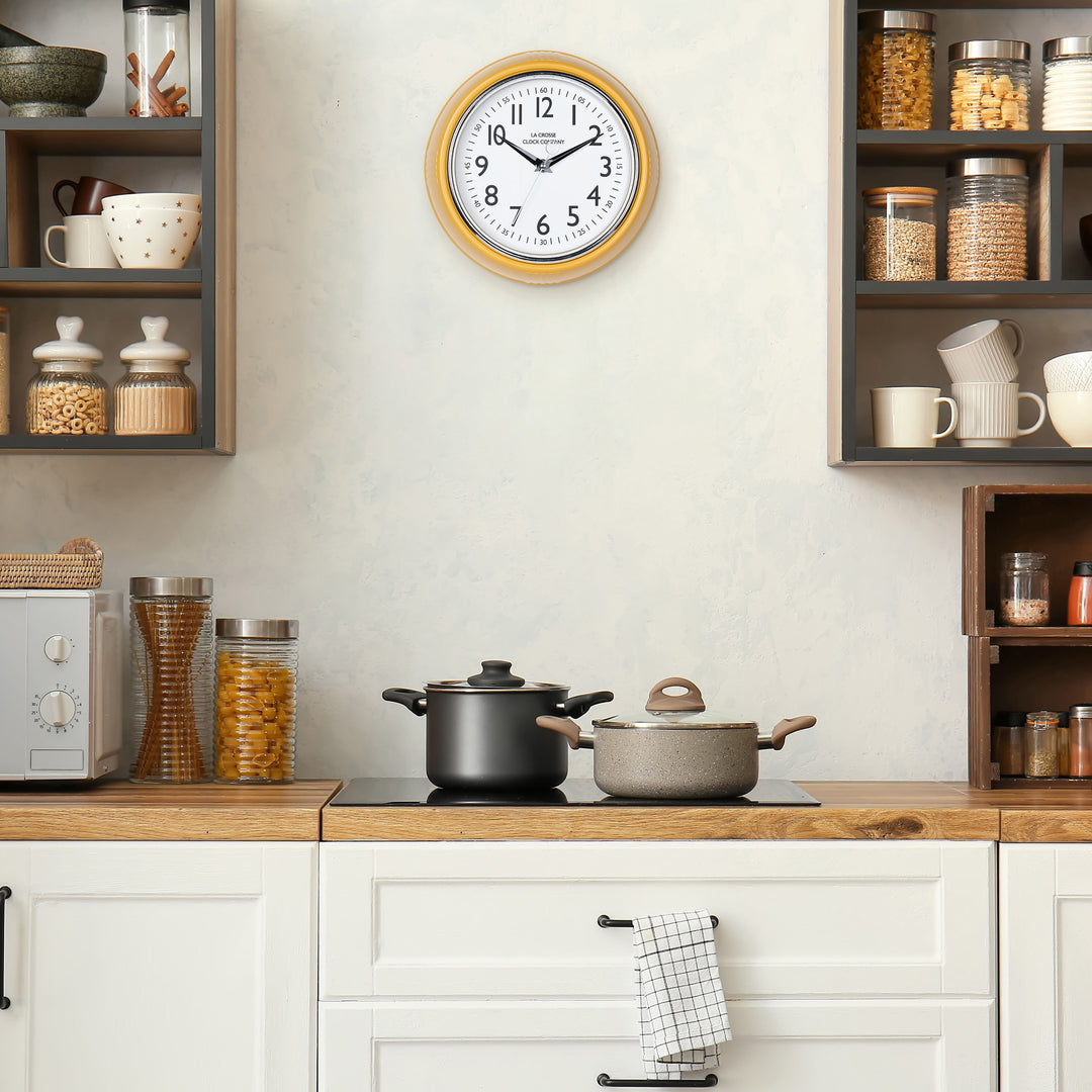 Lifestyle image of the 404-3024MY 9.5-inch Diner Clock – Mustard Yellow shown hanging on a cream kitchen wall above a stove top and cabinets. The clock features a retro-style mustard yellow frame with a white dial, chrome bezel, black numbers and minute markings, black metal hands, and a silver second hand.