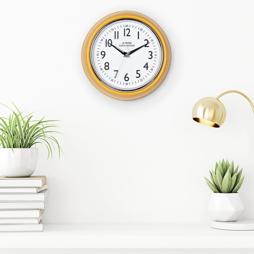 Lifestyle image of the 404-3024MY 9.5-inch Diner Clock – Mustard Yellow shown hanging on a white wall above a white shelf with plants, books, and a retro lamp. The clock features a retro-style mustard yellow frame with a white dial, chrome bezel, black numbers and minute markings, black metal hands, and a silver second hand.