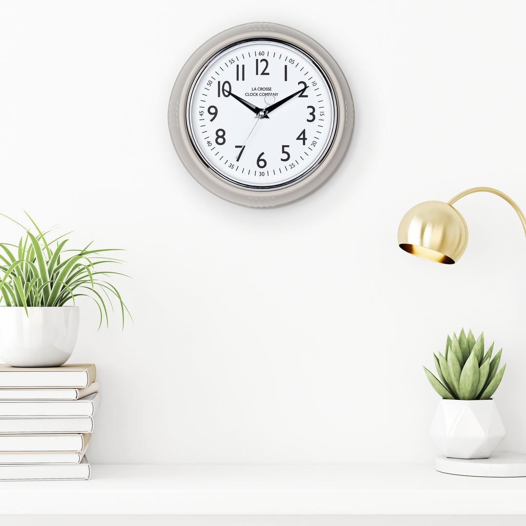 Lifestyle image of the 404-3024TP 9.5-inch Diner Clock – Taupe shown hanging on a white wall above a white shelf with plants, books, and a retro lamp. The clock features a retro-style taupe frame with a white dial, chrome bezel, black numbers and minute markings, black metal hands, and a silver second hand.