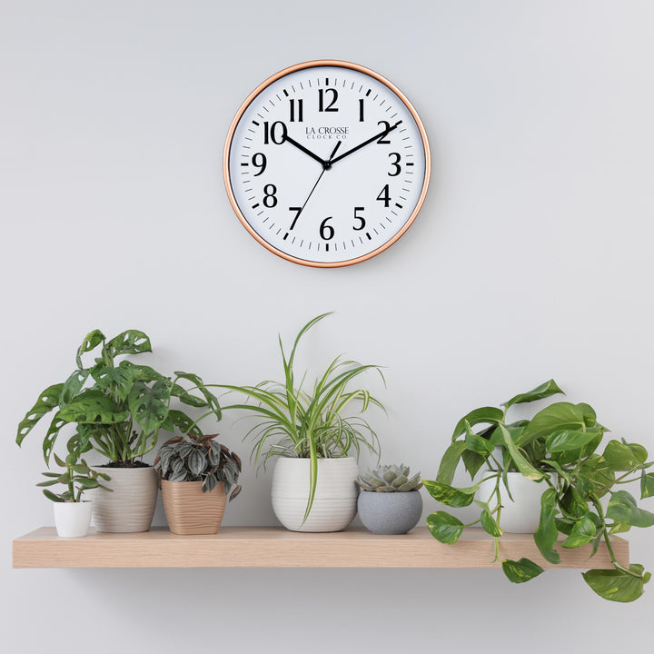 Lifestyle image of the 404-3025C 10-inch Basic Wall Clock - Copper hanging on a light grey wall above a pine shelf with multiple plants. The clock features a white dial, black numbers and markings, and hour, minute, and second hands, surrounded by a copper flush-mount frame.