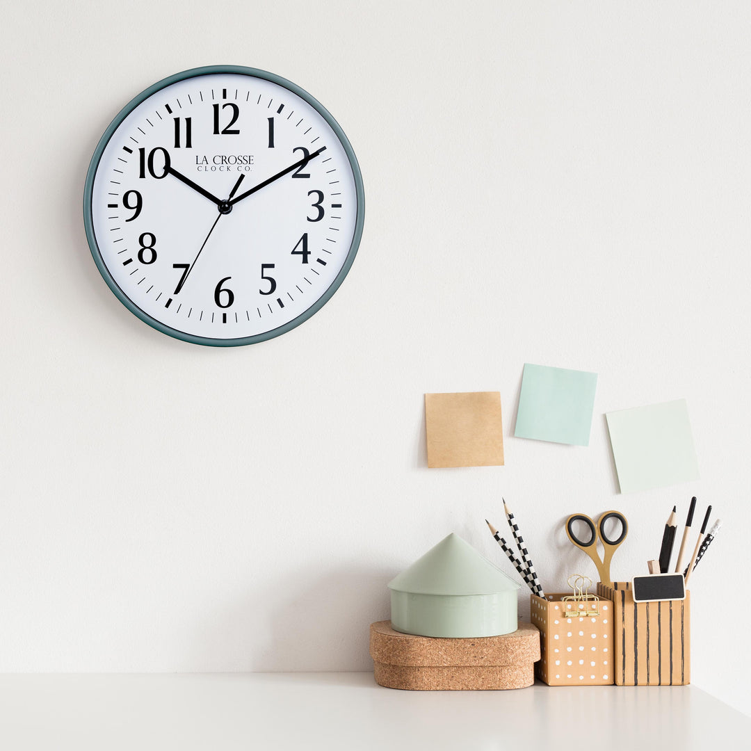 Lifestyle image of the 404-3025G 10-inch Basic Wall Clock - Green hanging on a white wall above a white desk with cork, wood, and pastel desk accessories. The clock features a white dial, black numbers and markings, and hour, minute, and second hands, surrounded by a green flush-mount frame.