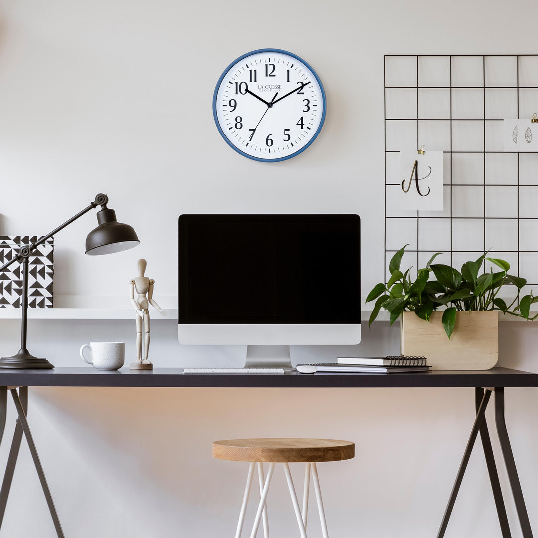 Lifestyle image of the 404-3025NB 10-inch Basic Wall Clock – Navy Blue, hanging on a light tan wall above a black desk. The desk has a computer, notebooks, a plant, a coffee mug, a black desk lamp, and a posable wood model. The clock features a white dial, black numbers, markings, hour, minute, and second hands surrounded by a Navy Blue flush-mount frame.