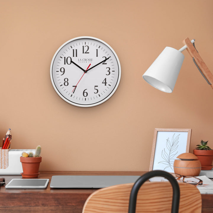 Lifestyle image of the 404-3025W 10-inch Basic Decor Wall Clock with a White frame, white dial with black numbers, hour, and minute hands, and a red second hand hanging on a neutral wall above a dark wood desk