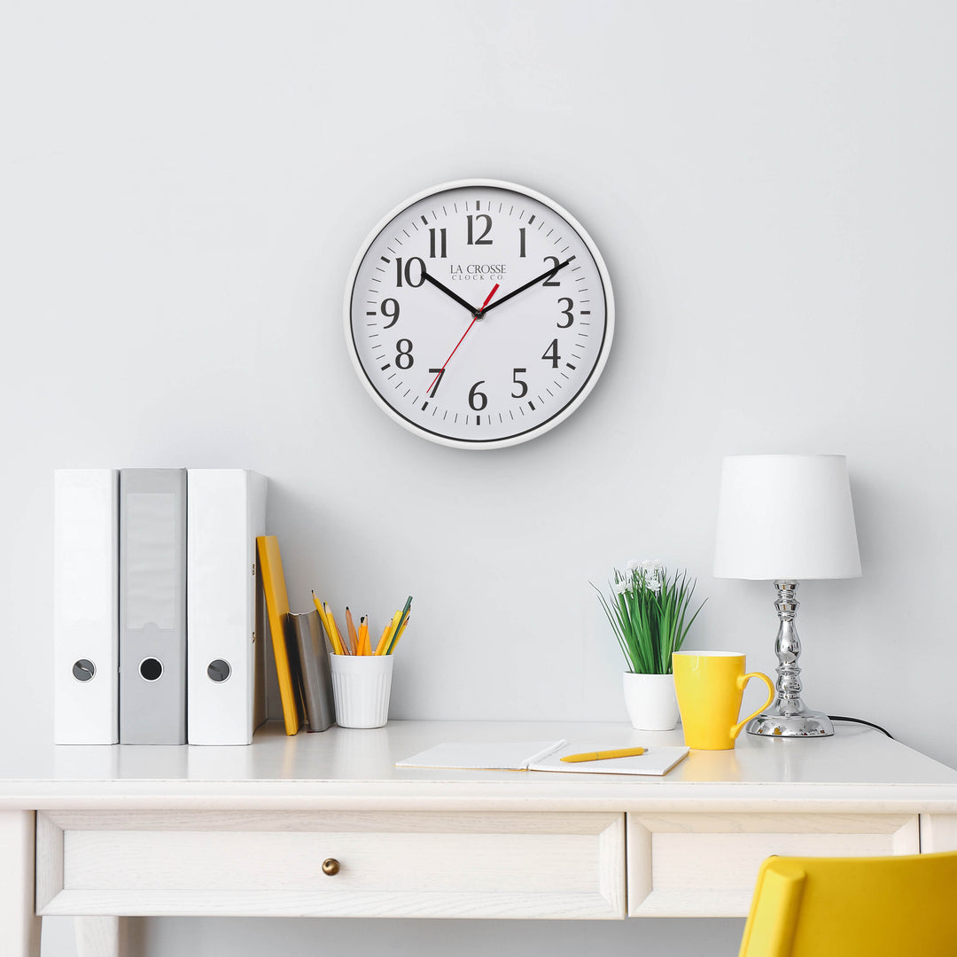 Lifestyle image of the 404-3025W 10-inch Basic Decor Wall Clock with a White frame, white dial with black numbers, hour, and minute hands, and a red second hand hanging on a white wall above a white desk with a lamp, notebook, pencil and desk accessories