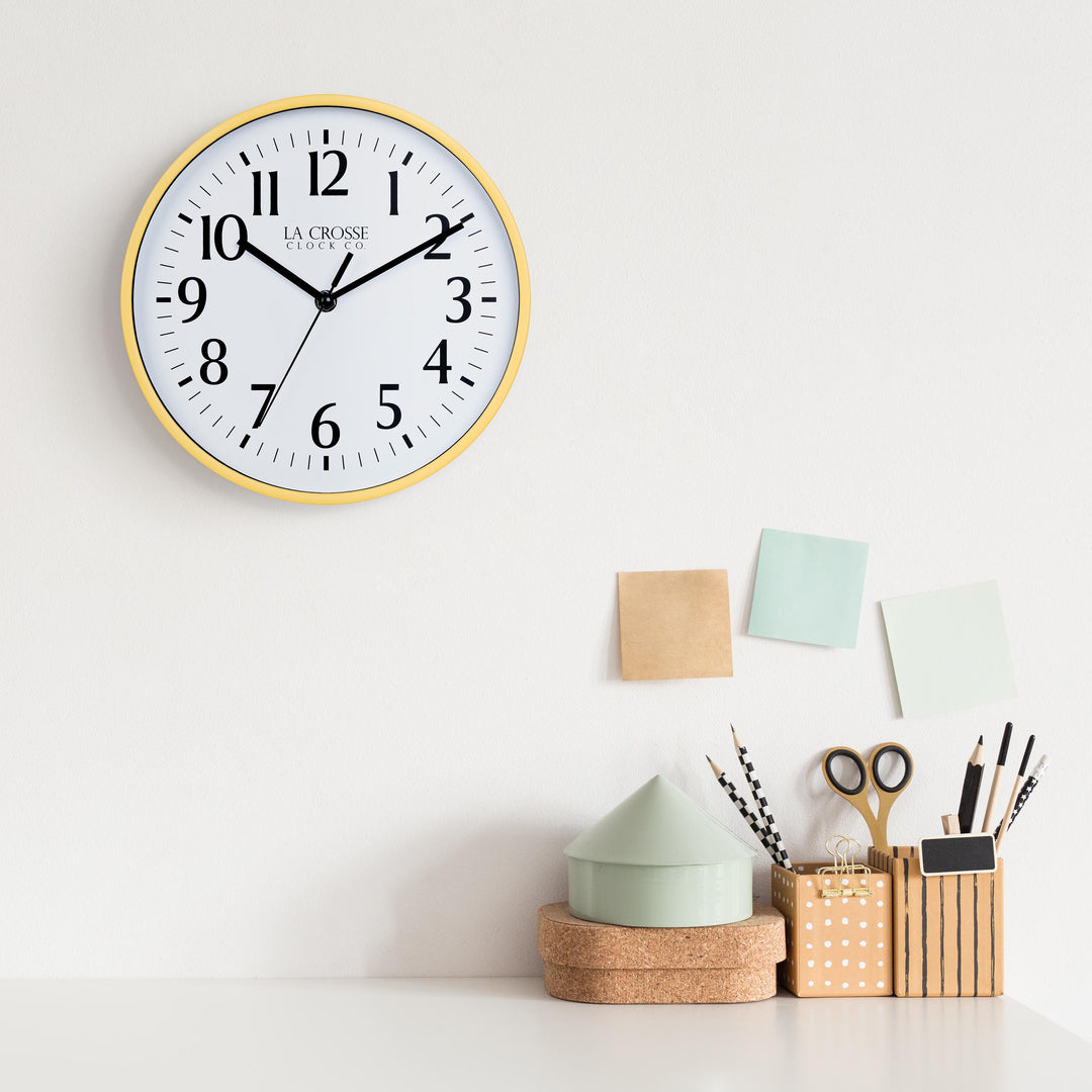 Lifestyle image of the 404-3025Y 10-inch Basic Wall Clock - Yellow hanging on a white wall above a white desk with cork, wood, and pastel desk accessories. The clock features a white dial, black numbers and markings, and hour, minute, and second hands, surrounded by a yellow flush-mount frame.