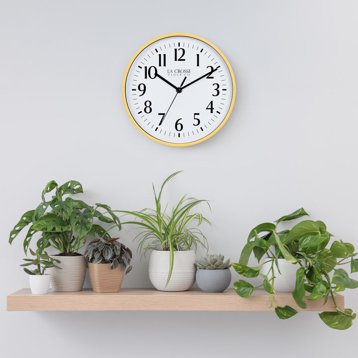 Lifestyle image of the 404-3025Y 10-inch Basic Wall Clock hanging on a light grey wall above a pine shelf with multiple plants. The clock features a white dial, black numbers and markings, and hour, minute, and second hands, surrounded by a yellow flush-mount frame.