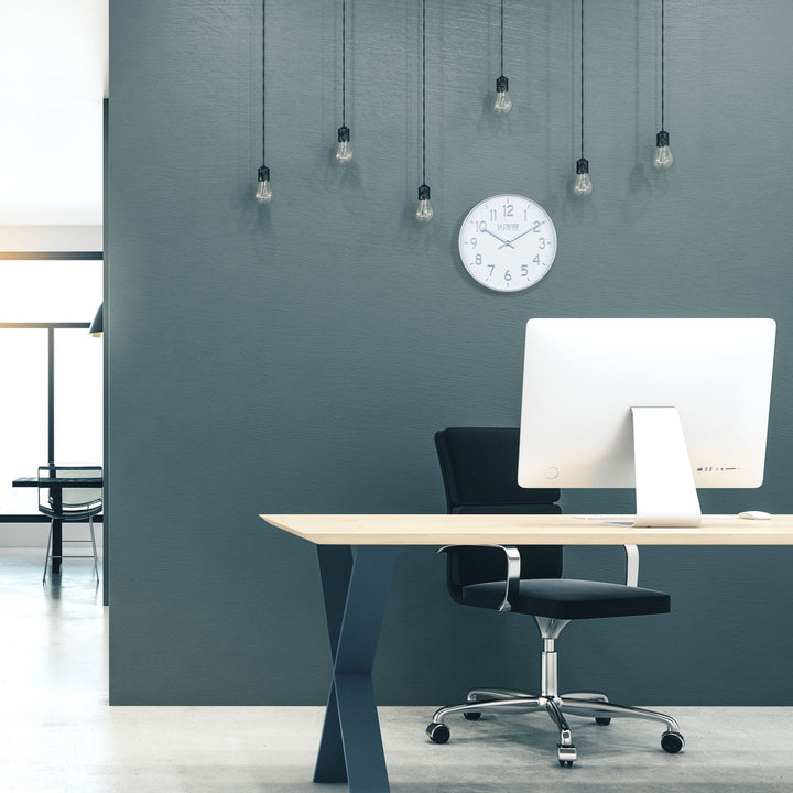 404-3027B Tovi wall clock with a white dial and silver Arabic numbers surrounded by a flush mount silver frame with matching silver hour and minute hands hanging on a wall in a modern office setting of dark gray walls light gray flooring and a light wood top desk with a laptop and office chair