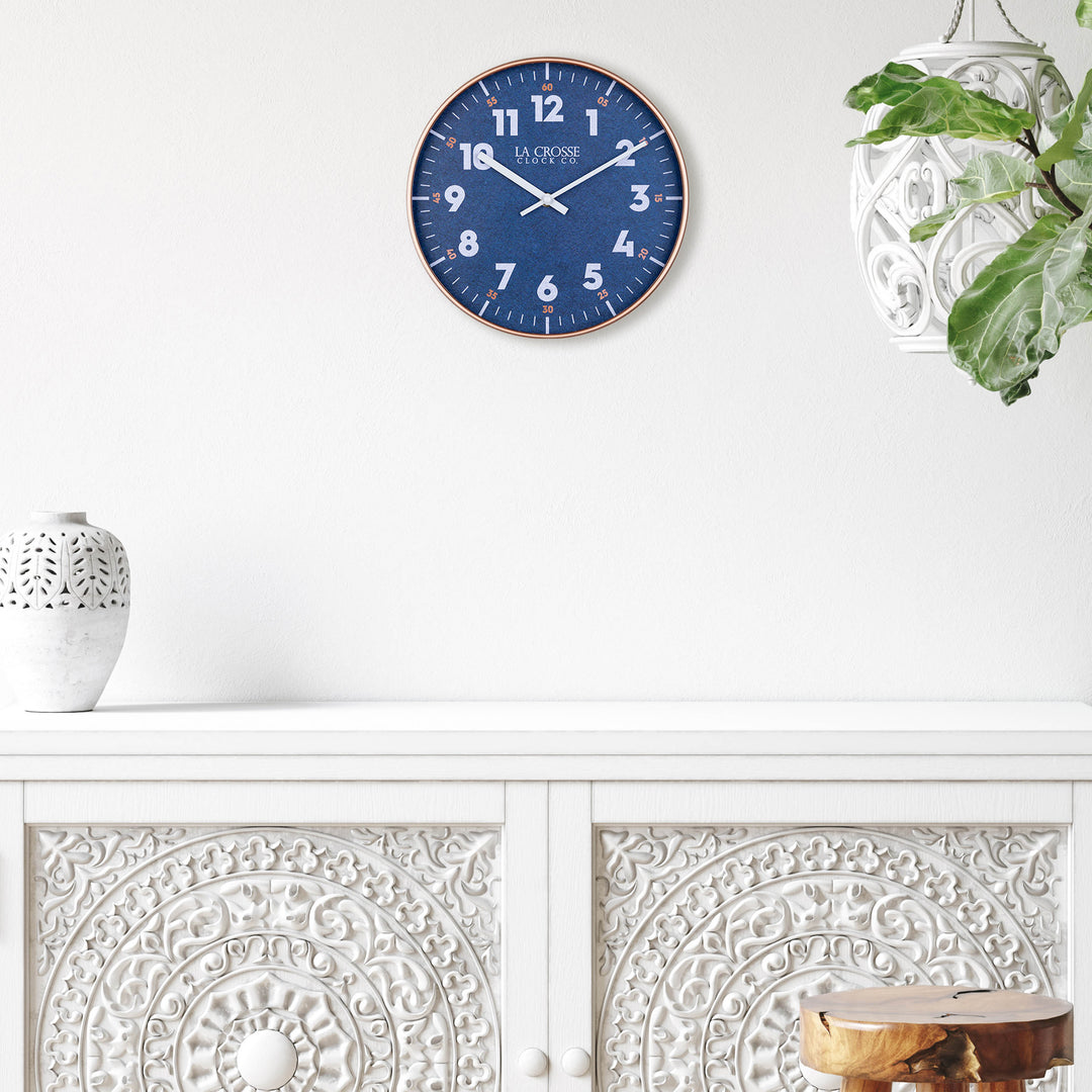 Lifestyle image of the 404-3027C Portside eleven inch navy blue wall clock with white numbers, orange time markings, and a copper case featuring white hour and minute hands over a buffet side table of white with a carved white planter and vase