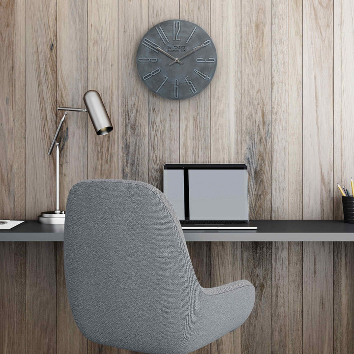 404-3028B Cormac 11 inch open face molded dark gray wall clock with numbers in relief and silver metal hour and minute hands hanging on a paneled wall with a gray office chair at a desk with a laptop and silver office lamp