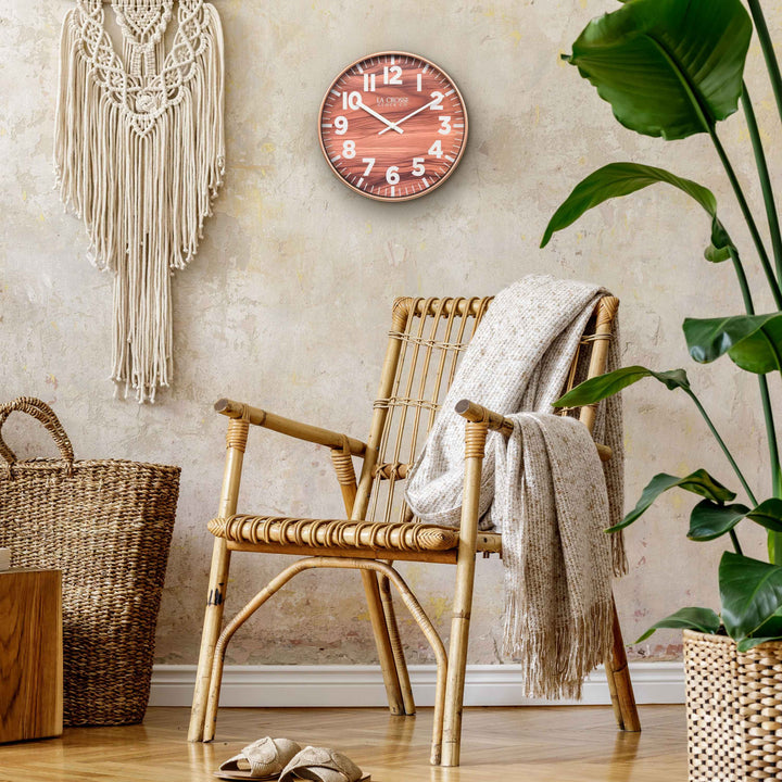 Lifestyle image of the 10.8-inch Atwood Wall Clock hanging on a tan textured wall above a wicker chair with Bohemian style decor. The clock features a copper finish frame, a wood design dial, white numbers, and white metal hour and minute hands.