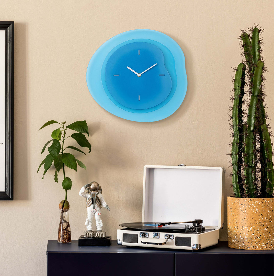 Lifestyle image of the 404-3039BL 15.2-inch Drift Wall Clock – Blue, shown hanging on a tan wall above a black side table with a vintage turntable, cactus, a plant, and a statue of an astronaut. The clock features an open face multi-layered design with drifting contours, metal hands, and raised number markings in white.