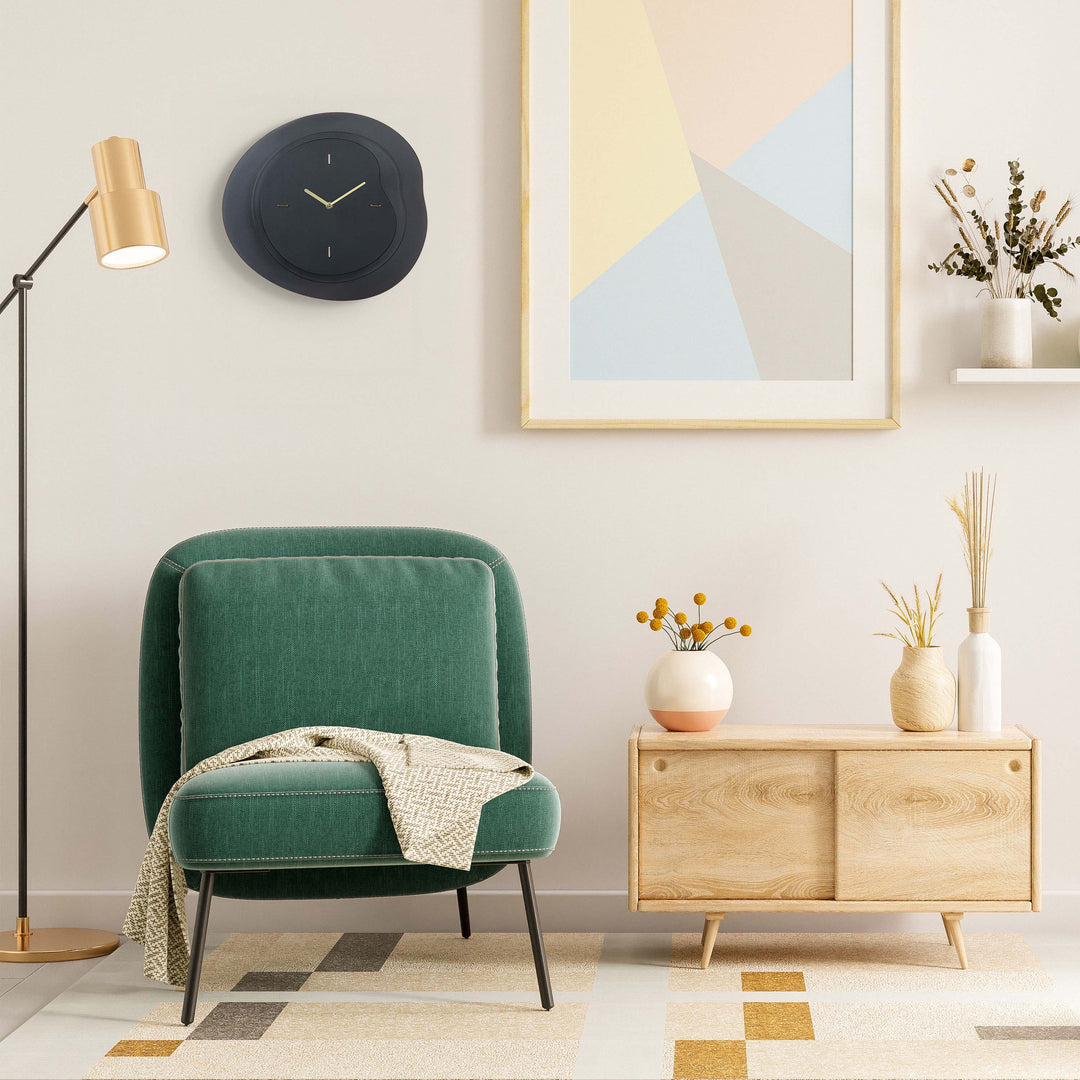 Lifestyle image of the 404-3039B 15.2-inch Drift Wall Clock – Black, shown hanging on a tan wall above an emerald green chenille chair, retro brass floor lamp, and light wood side cabinet. The clock features an open face multi-layered design with drifting contours, metal hands, and raised number markings in a brass finish.