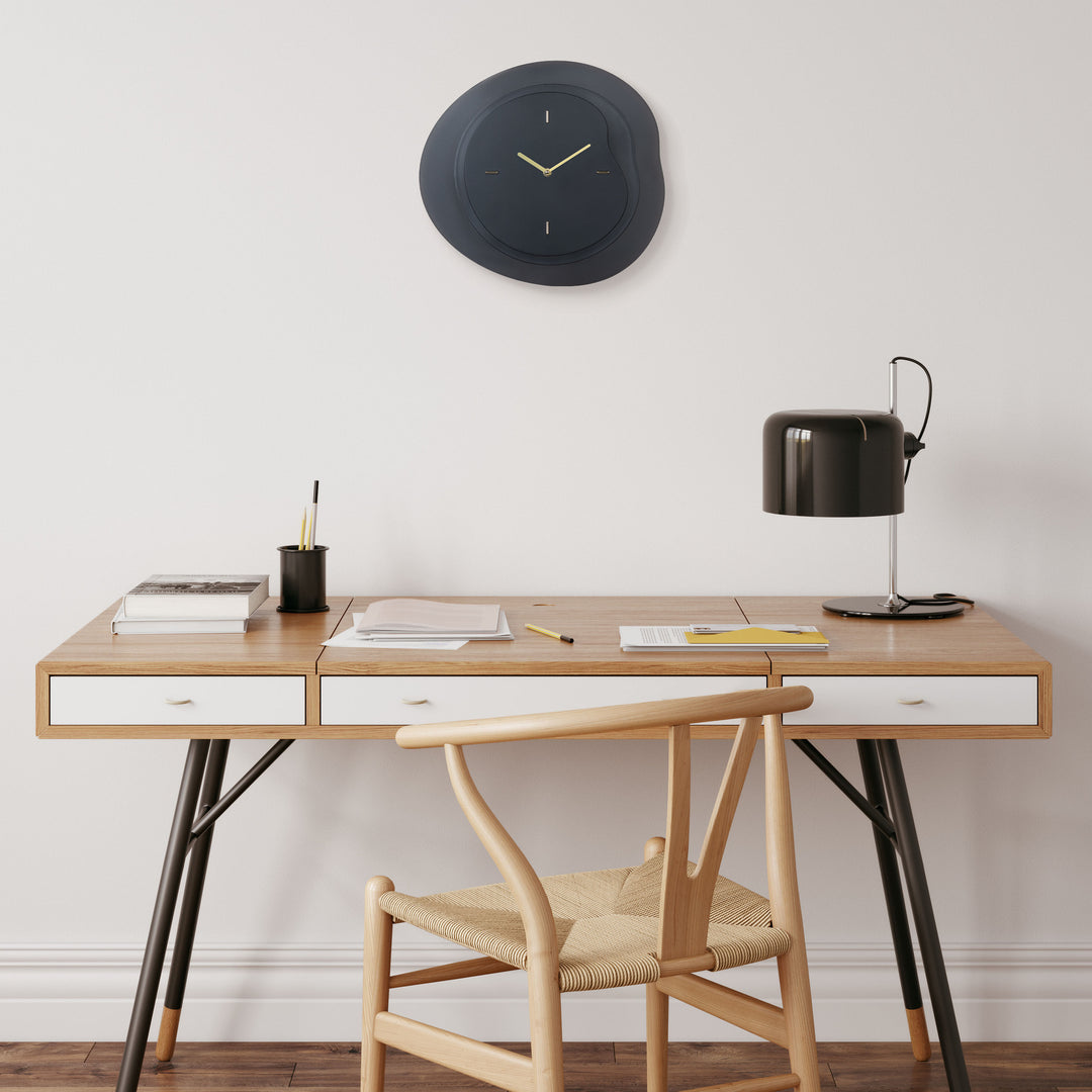 Lifestyle image of the 404-3039B 15.2-inch Drift Wall Clock – Black, shown hanging on a light wall above a modern black and wood desk. The clock features an open face multi-layered design with drifting contours, metal hands, and raised number markings in a brass finish.