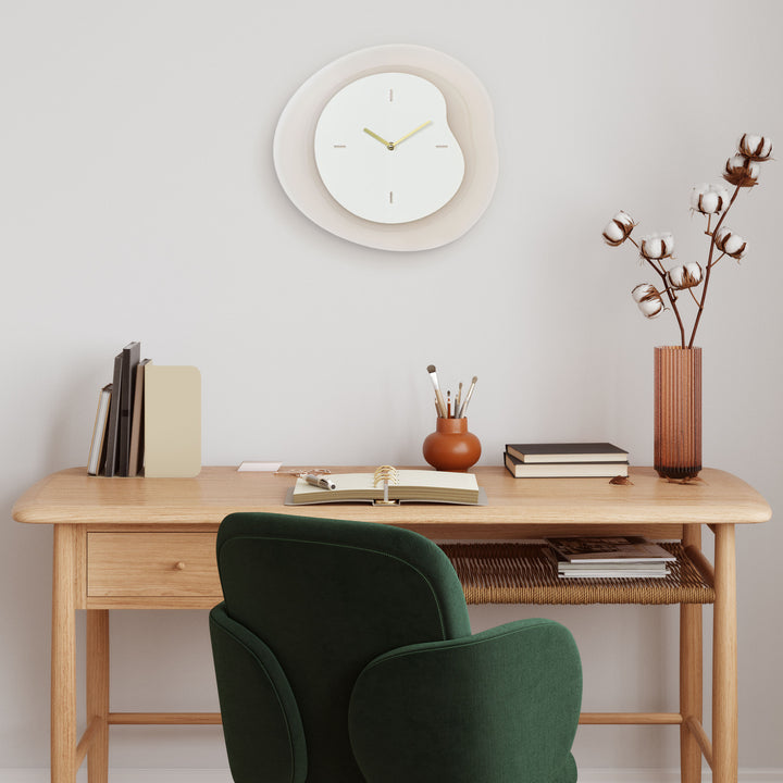 Lifestyle image of the 404-3039T 15.2-inch Drift Wall Clock – Tan, shown hanging on a cream wall in a Transitional Style home office with a light wood desk and green velvet chair. The clock features an open face multi-layered design with drifting contours, metal hands, and raised number markings in a brass finish.