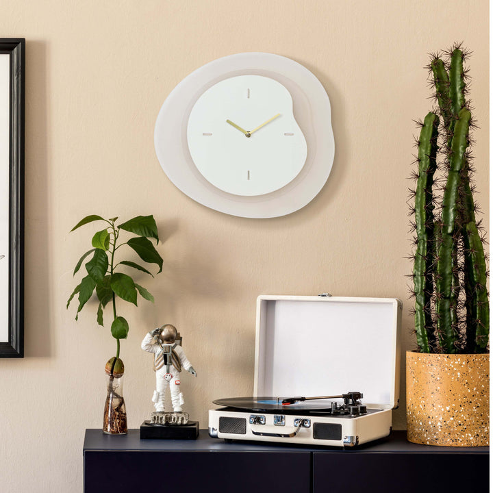 Lifestyle image of the 404-3039T 15.2-inch Drift Wall Clock – Tan, shown hanging on a tan wall above a black side table with a vintage turntable, cactus, a plant, and a statue of an astronaut. The clock features an open face multi-layered design with drifting contours, metal hands, and raised number markings in a brass finish.
