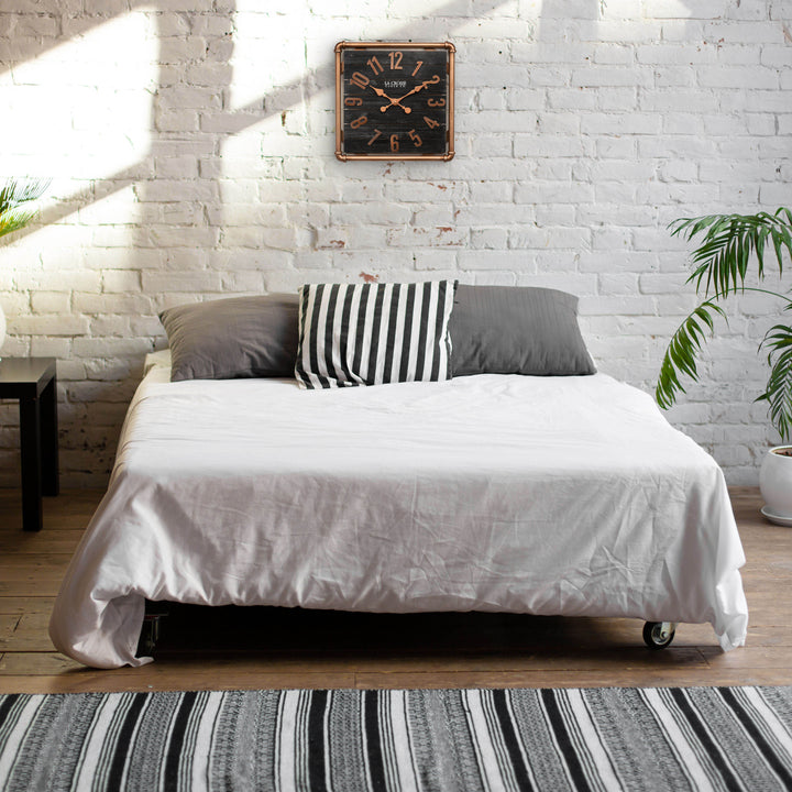 Lifestyle image of the 404-3412D 16-inch Foundry Metal &amp; Wood Wall Clock - Dark, shown hanging on a white-washed brick wall above a bed. The clock features a square metal pipe frame in a copper finish, a black-stained wood slat clock face with copper-finished metal numbers and hands.